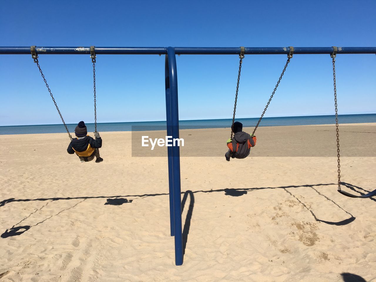 Rear view of children playing on swing at beach against clear blue sky during sunny day