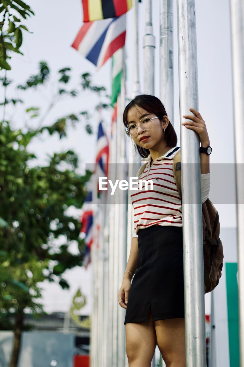 Portrait of young woman standing by pole against sky