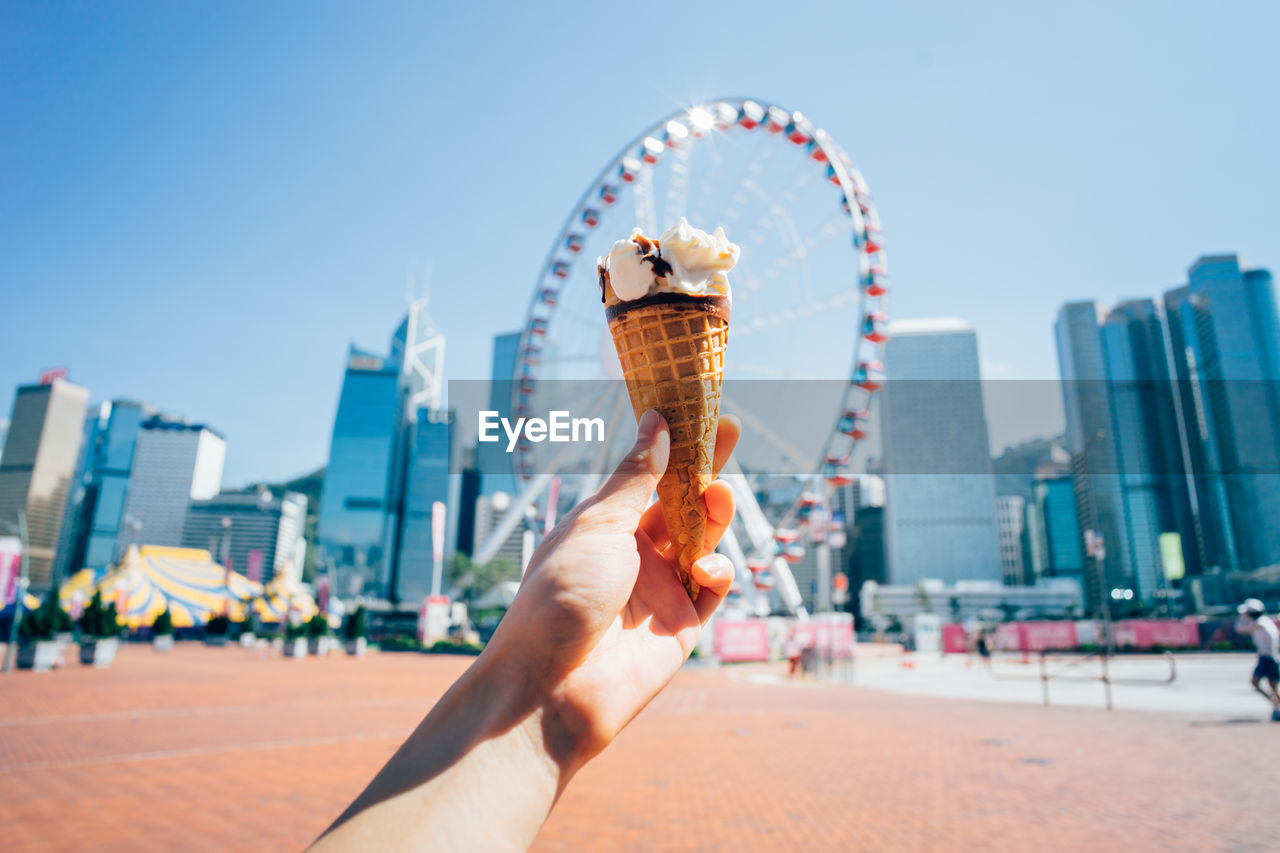 Cropped image of hand holding ice cream against ferris wheel in city