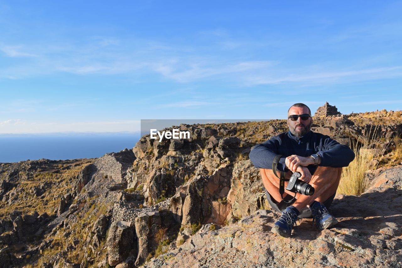 Portrait of mature man holding camera while sitting on mountain against blue sky