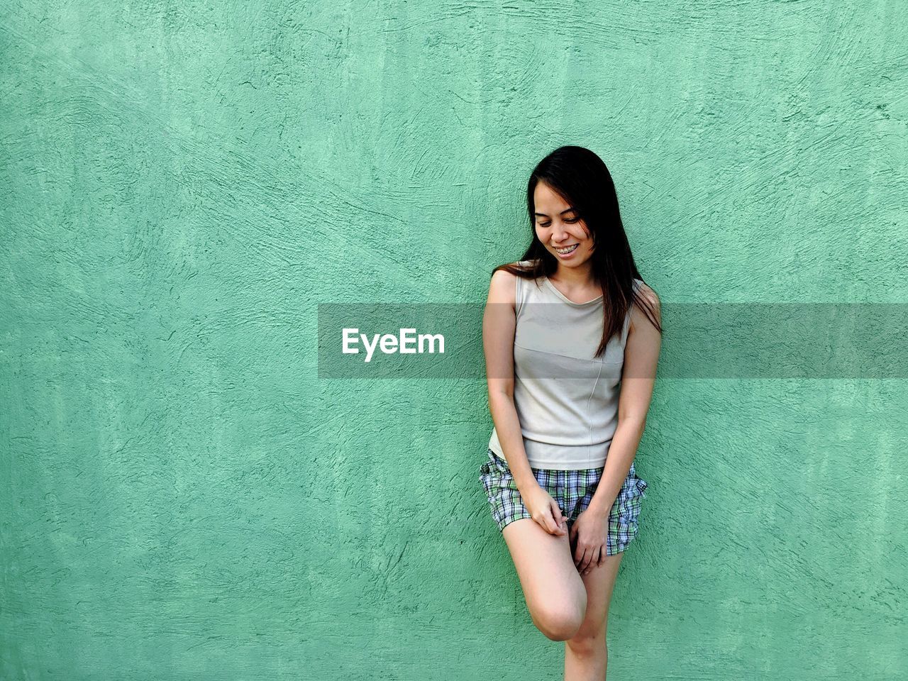 Smiling young woman looking down while standing against wall