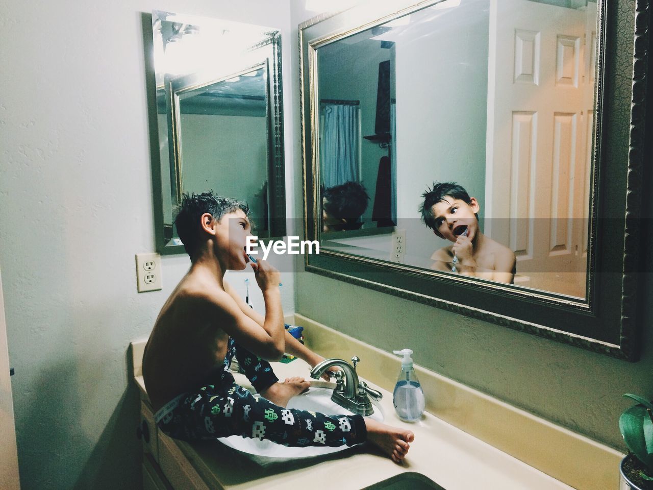 Cute shirtless boy brushing teeth in front of mirror while sitting on sink