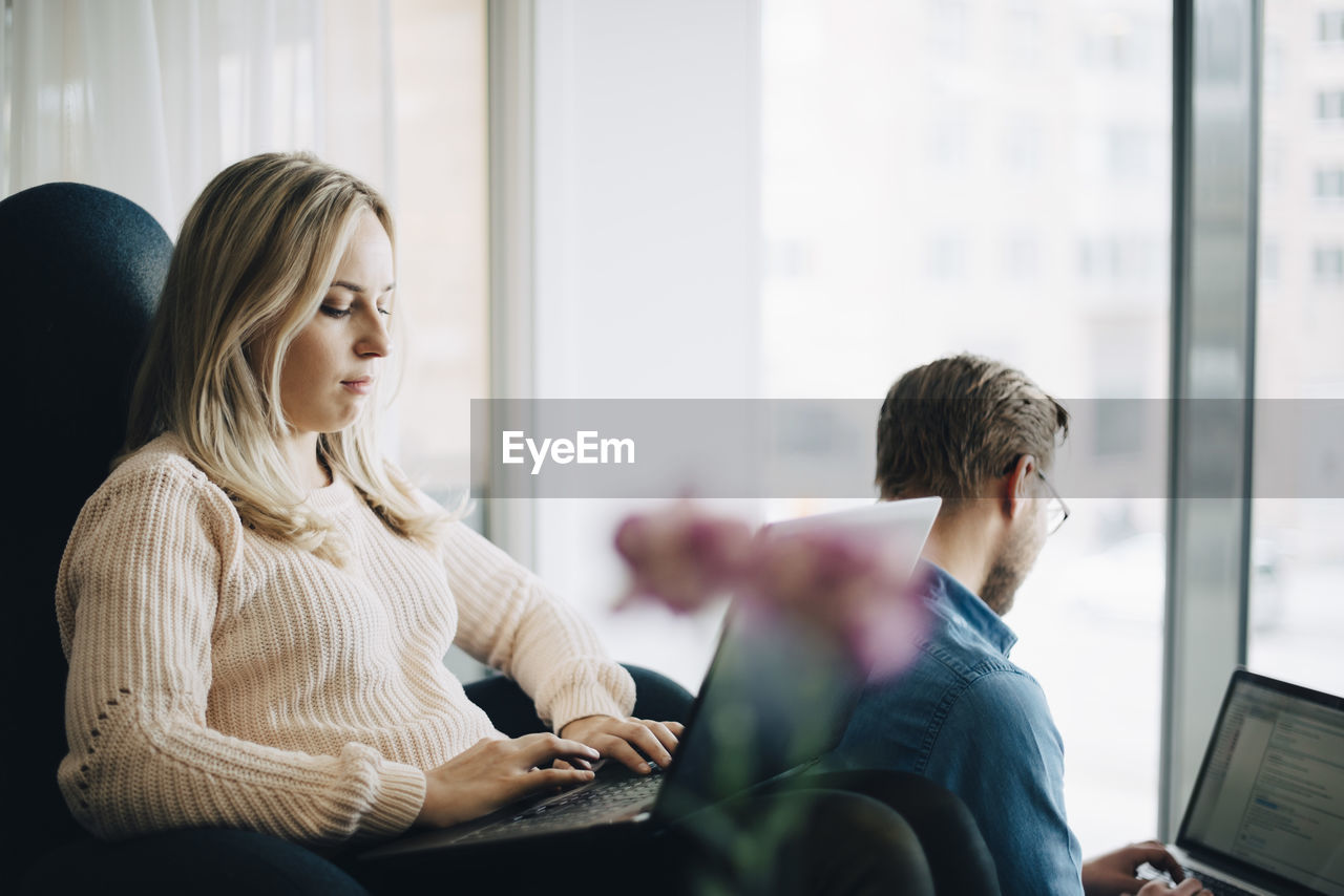 Young businesswoman sitting on chair using laptop by businessman at office cafeteria