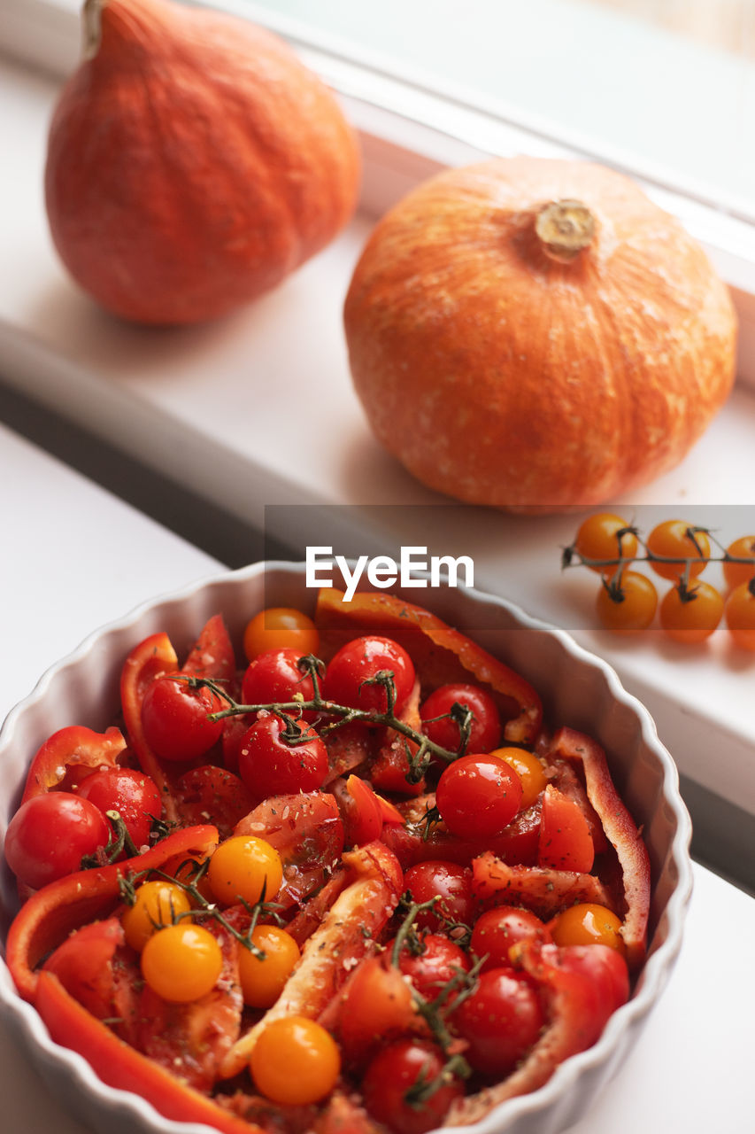 Baked various tomatos standing on the table against windowsill with autumn vegetables.