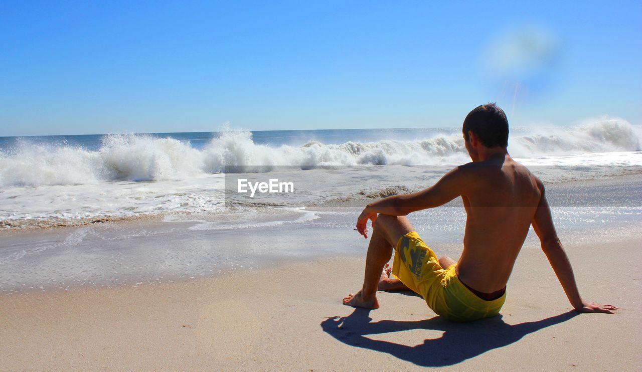 Rear view full length of shirtless man sitting at beach against waves