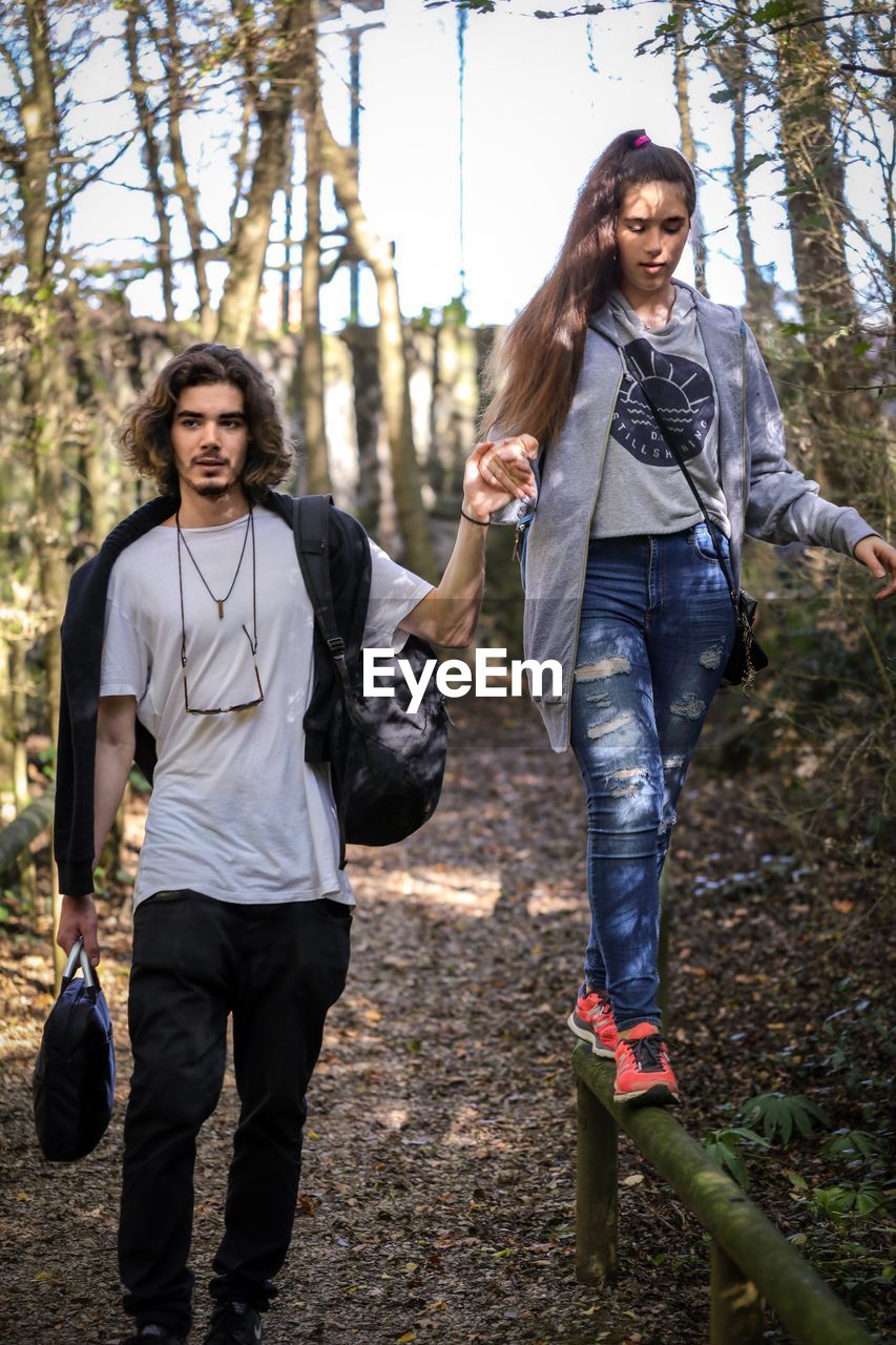 Young man assisting sister walking on railing in forest