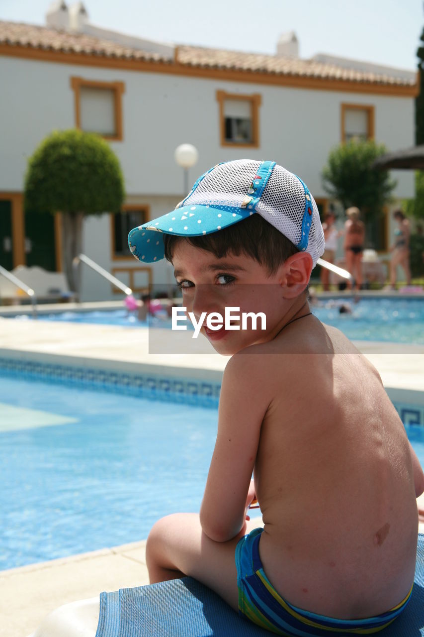 Portrait of shirtless boy sitting on pool side on sunny day