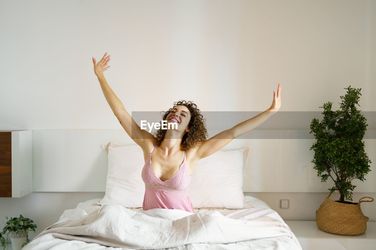 portrait of smiling young woman with arms raised standing against wall