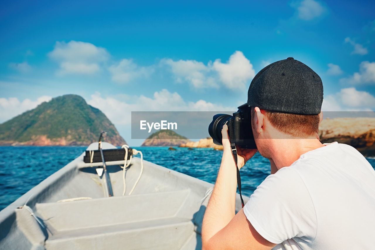 Side view of man photographing while traveling in boat on sea against blue sky