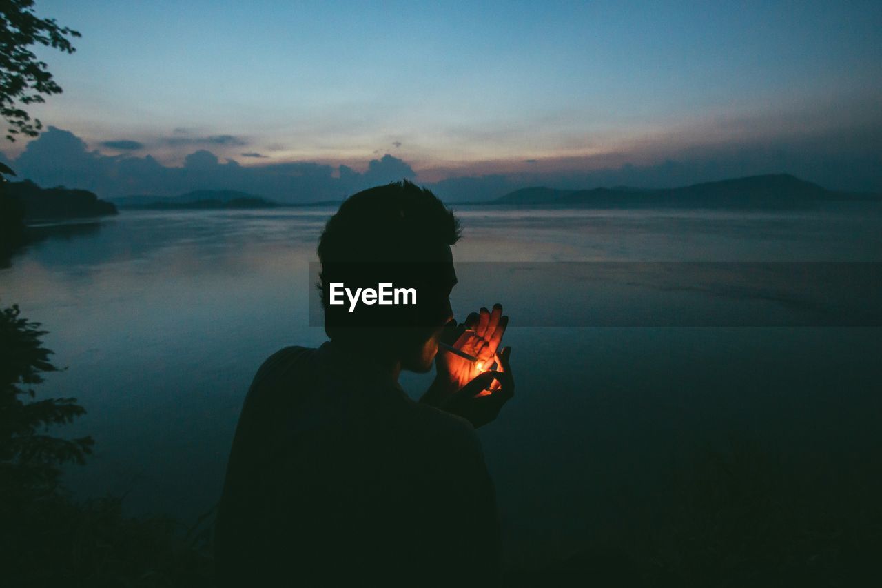 Man lighting cigarette while sitting in front of river