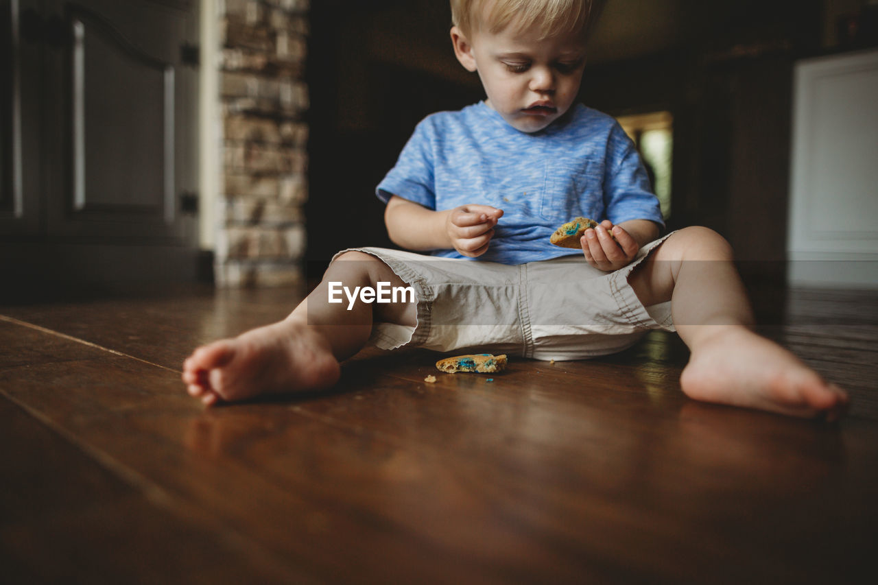 Cute baby boy eating cookie while sitting on floor at home