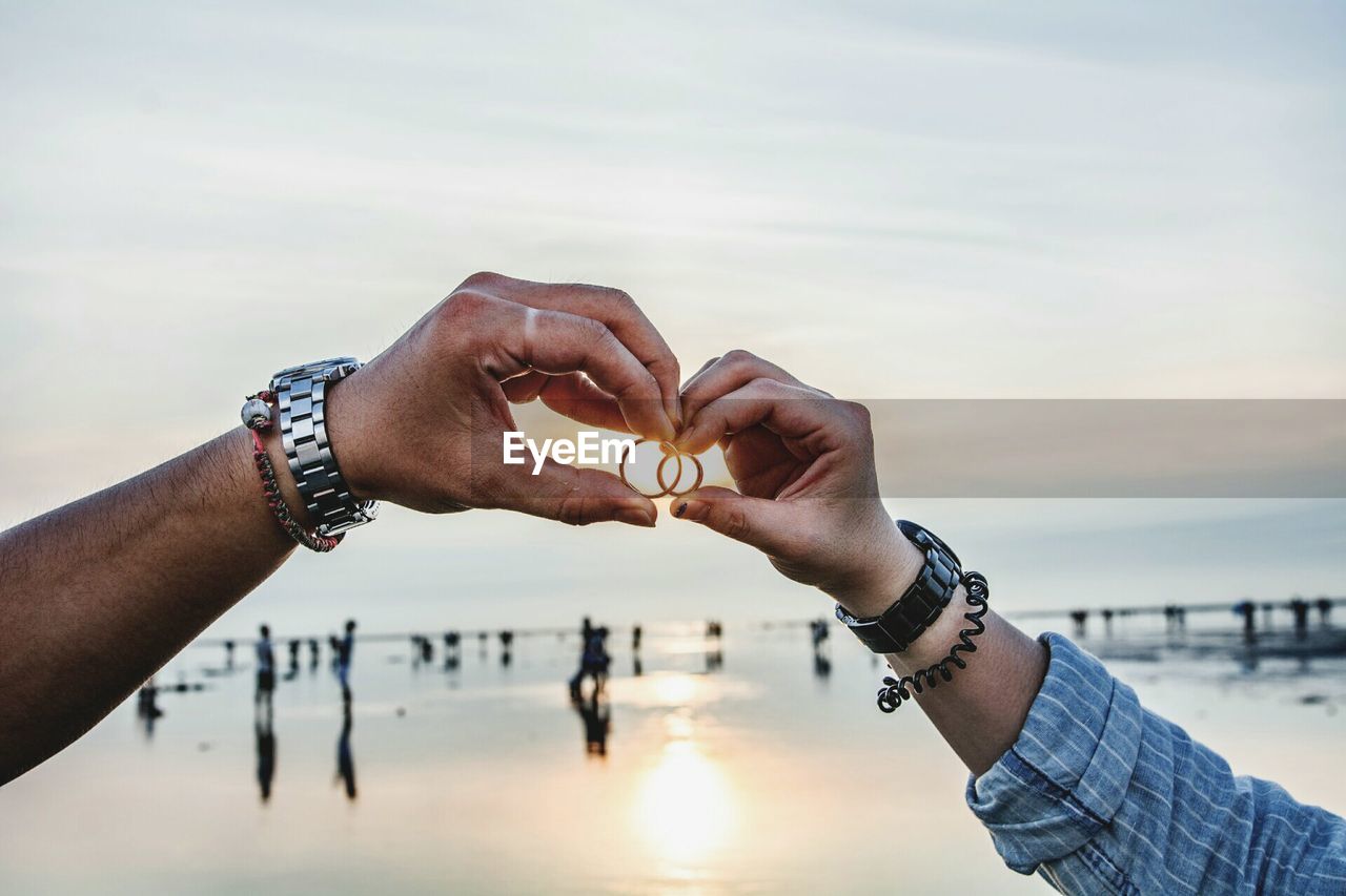 Cropped image of couple holding rings at beach against sky