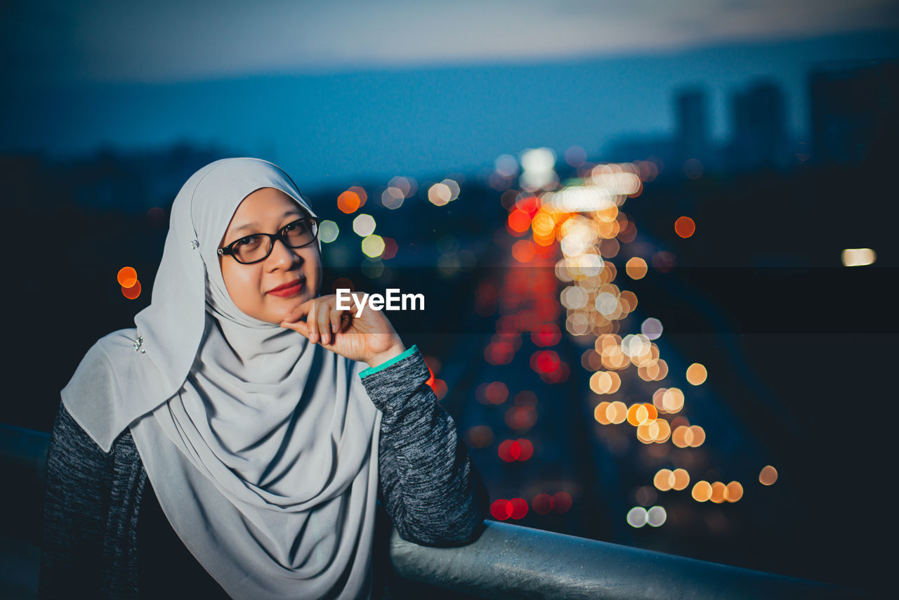 Portrait of woman wearing hijab by railing against illuminated city