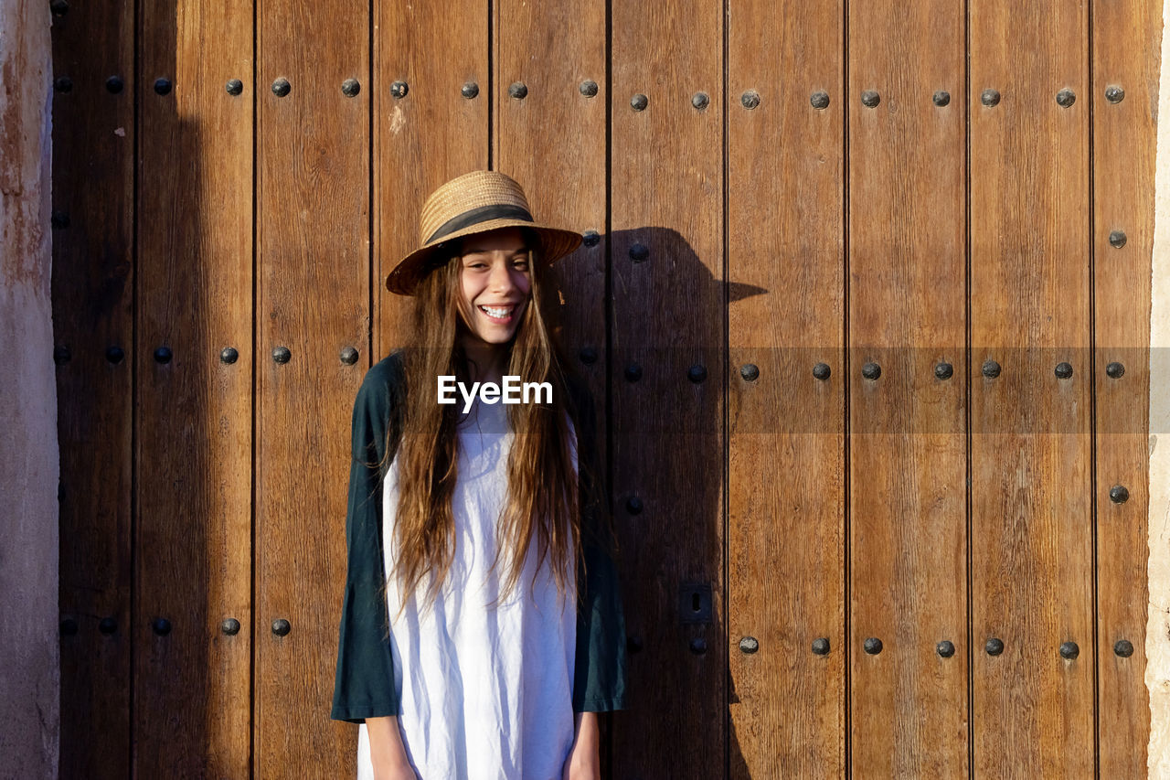 Portrait of young woman wearing hat standing against wooden wall