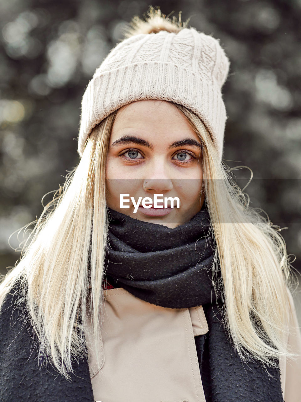 Young sincere blond female in knitted hat and warm clothes looking at camera in back lit