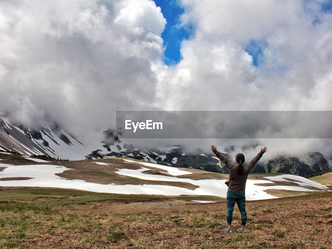 Rear view of hiker standing with arms raised against clouds covering mountains