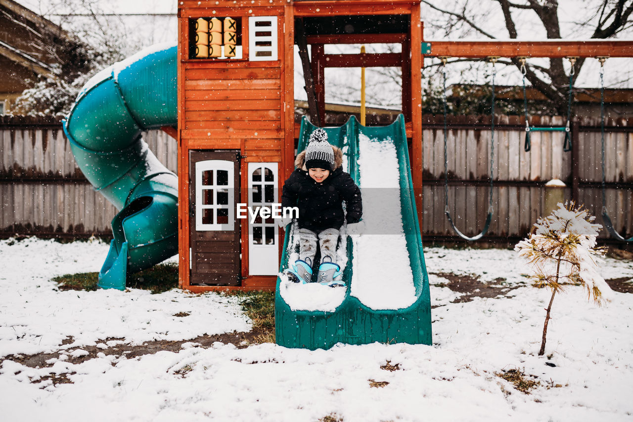 Young boy sliding on snow covered swingset in backyard at home