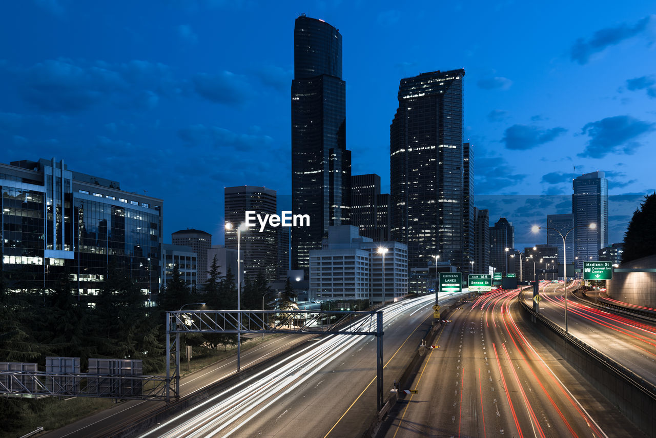 Light trails on road amidst buildings against sky at night