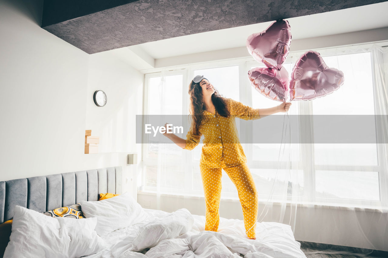 full length of woman with arms raised standing on bed at home