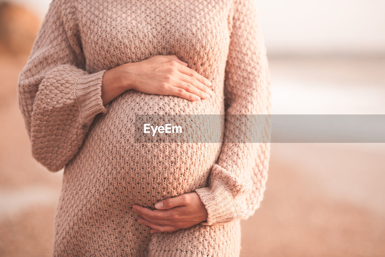 Pregnant woman wearing cozy knitted sweater holding tummy with hands outdoors close up. 