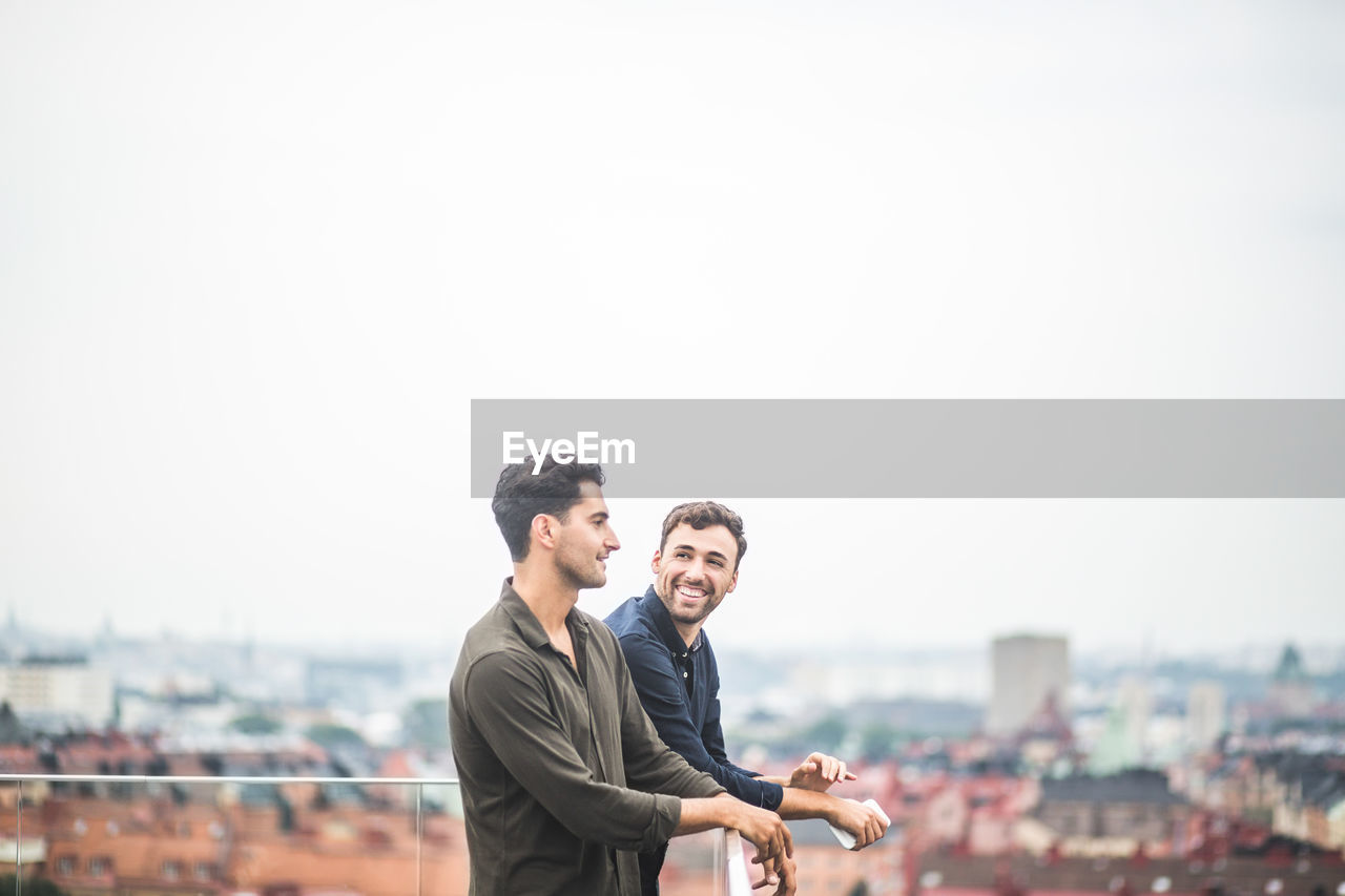 Smiling man looking at male friend while leaning over glass railing on rooftop against sky