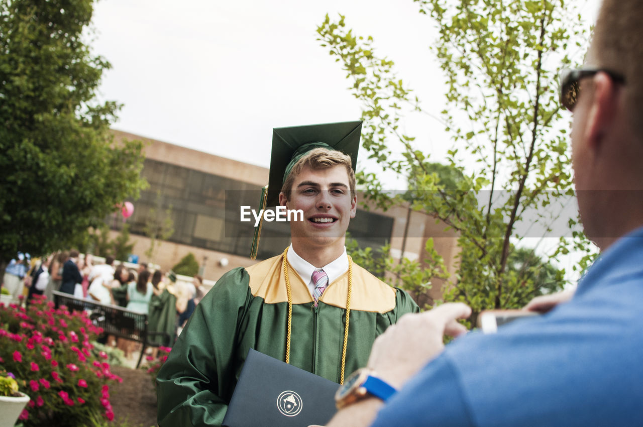 Father photographing male graduate student in campus at graduation ceremony