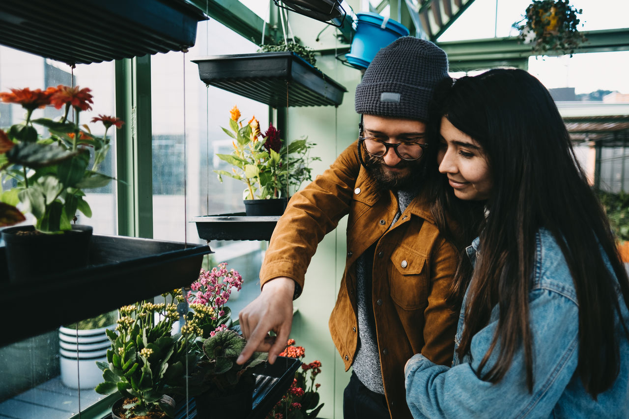 Content stylish couple embracing in greenhouse while standing near shelf and picking blooming potted flower
