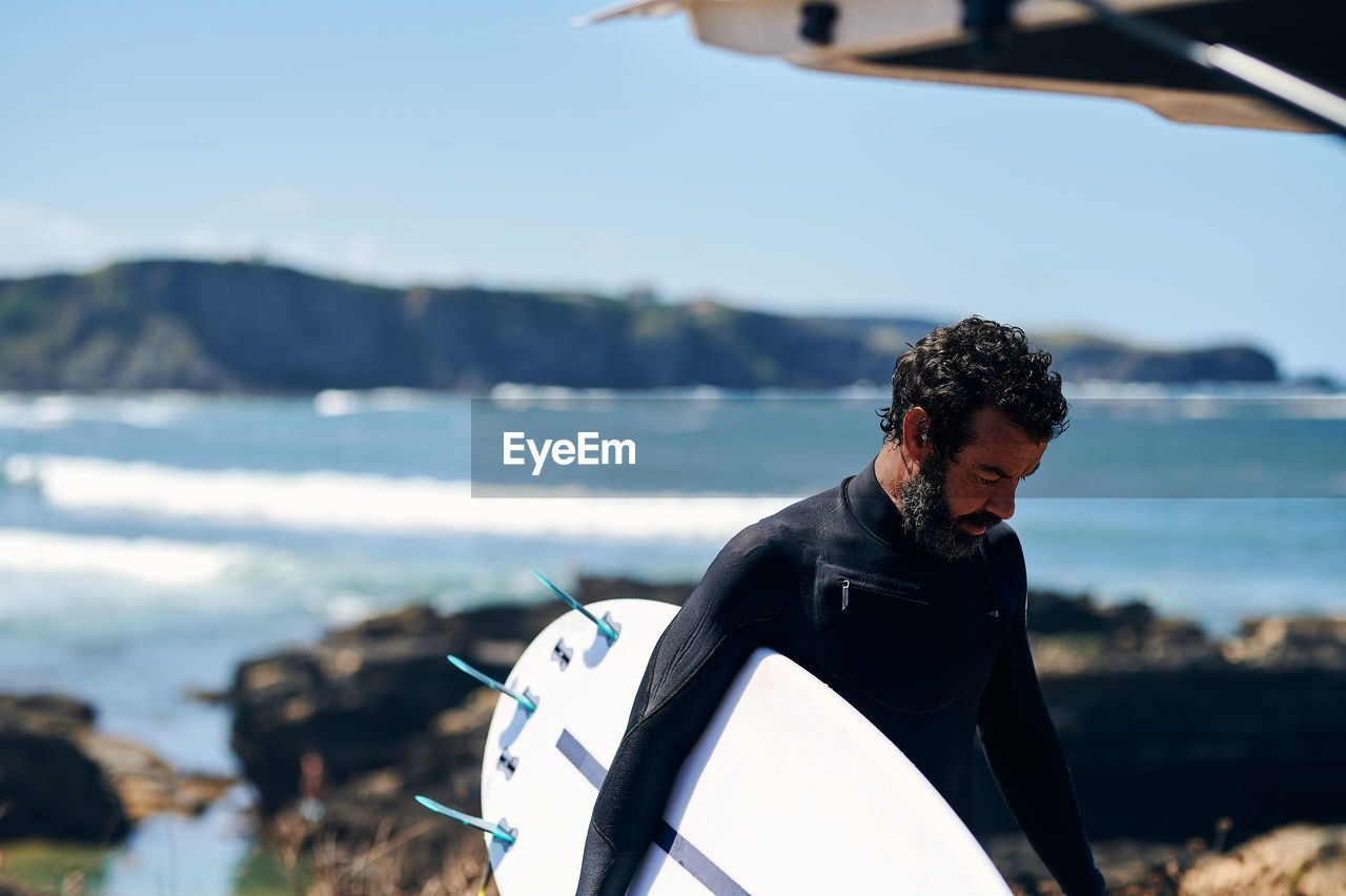 Adult man in wetsuit with surfboard smiling happily standing on seashore against hills