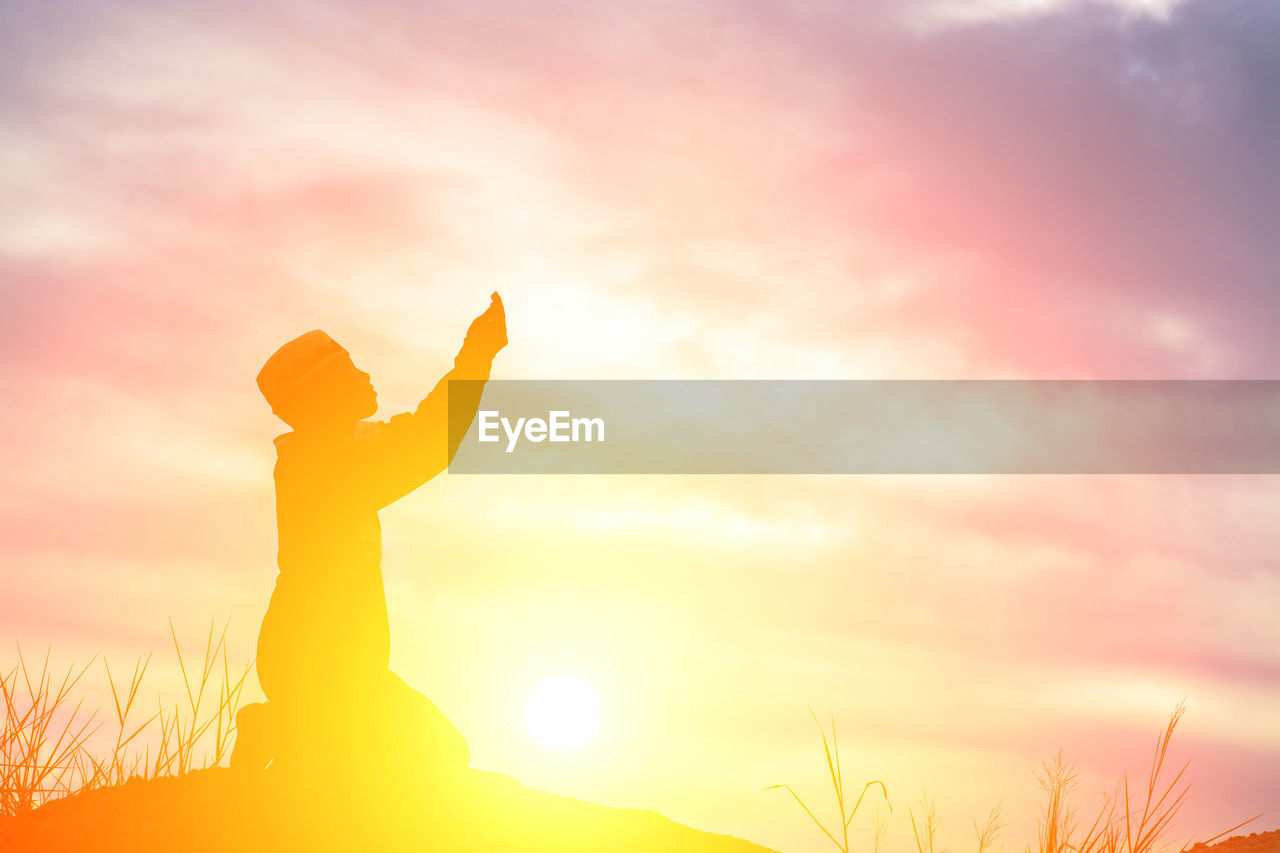 Silhouette boy kneeling on field against sky during sunset