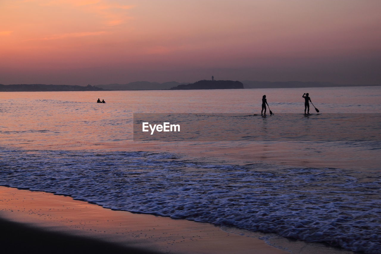 Silhouette man and woman paddleboarding on sea against sky during sunset