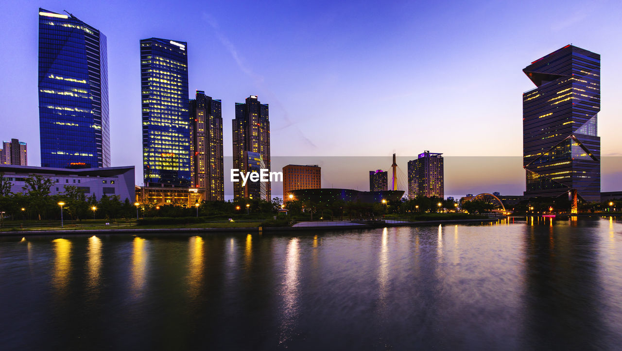 Illuminated modern buildings by river against sky in city