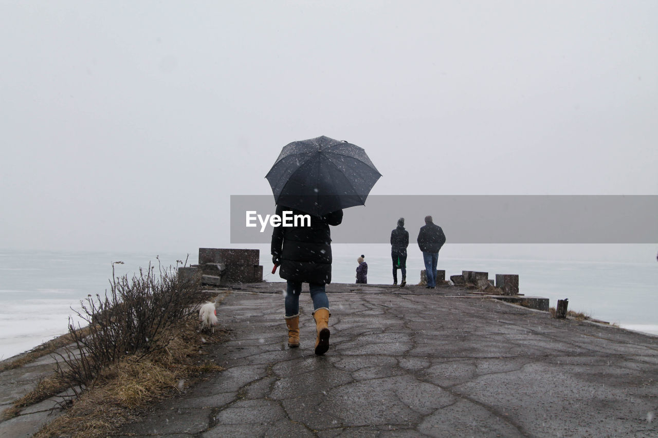 Rear view of people walking on footpath leading towards sea against sky