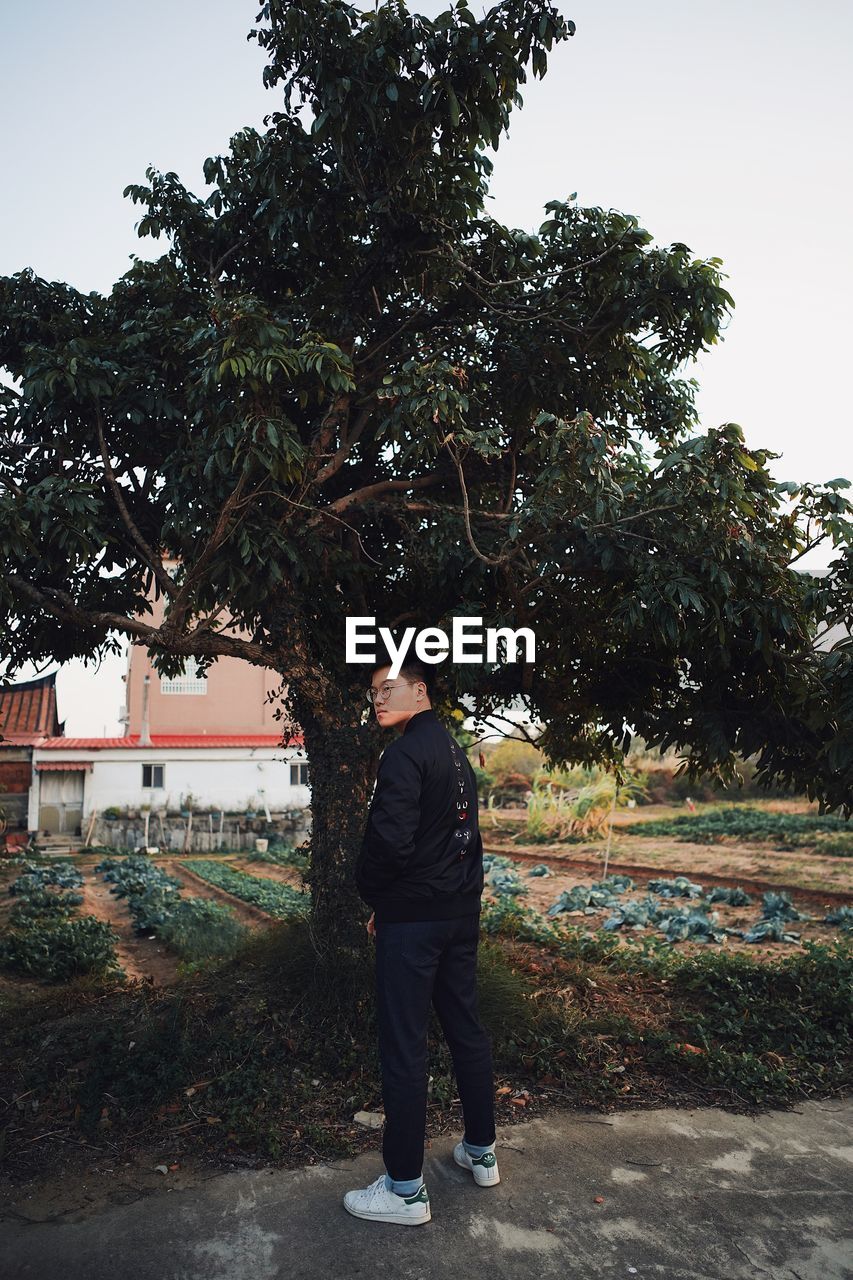 MAN STANDING ON FOOTPATH BY TREE AGAINST SKY