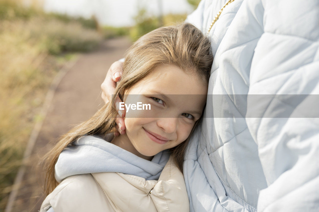 Crop mother in outerwear pressing head of cheerful daughter to chest while spending weekend day in park