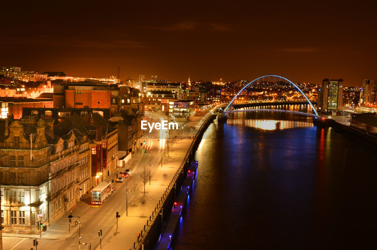 HIGH ANGLE VIEW OF ILLUMINATED BRIDGE OVER RIVER AGAINST BUILDINGS AT NIGHT