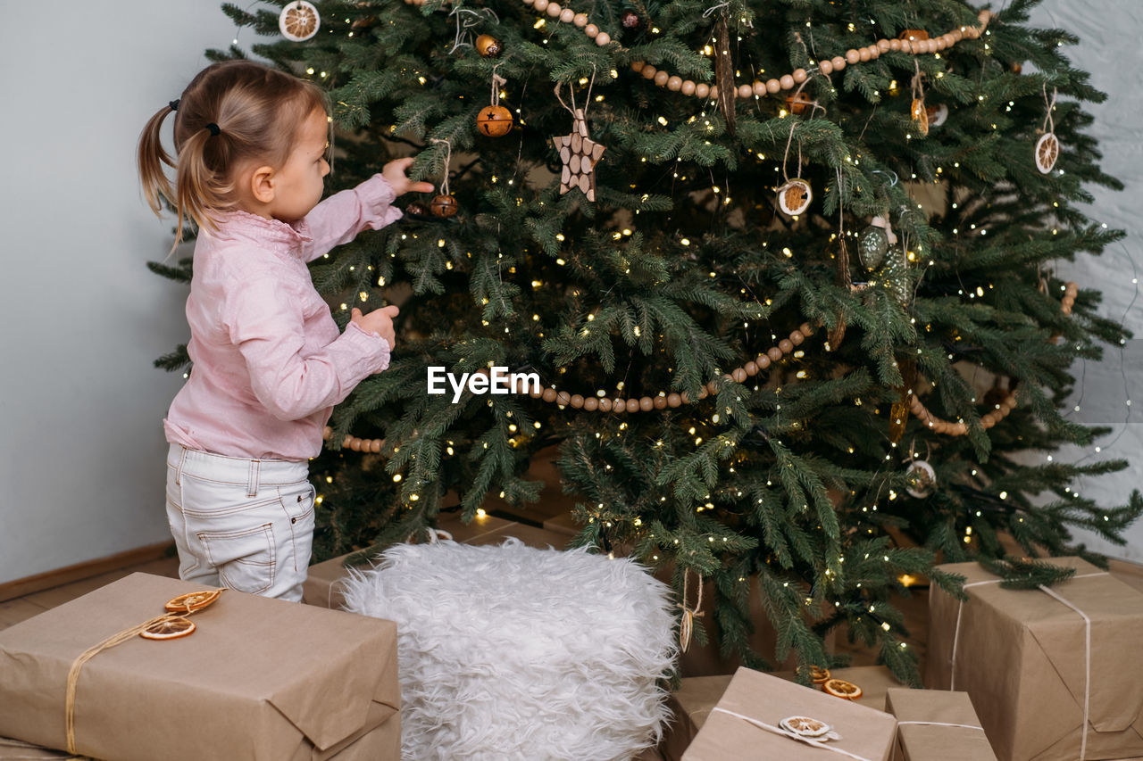 Cute toddler child waiting for presents at home after decorating christmas tree in eco style with