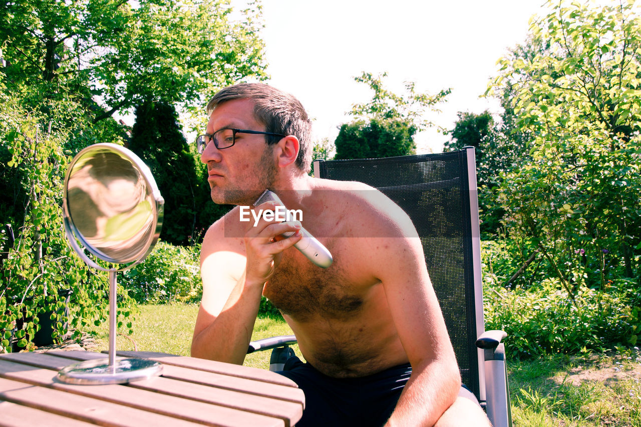 Shirtless man trimming beard in yard