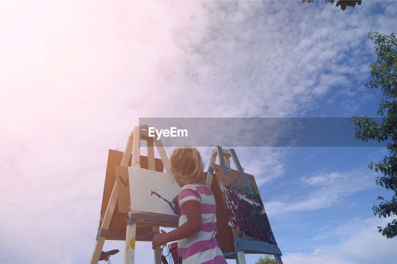 Low angle view of girl painting while standing against sky