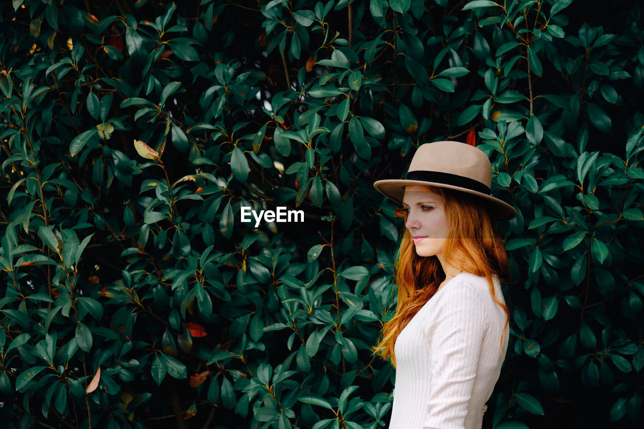 Attractive young long haired ginger female in stylish hat looking away while standing against green bushes in summer garden