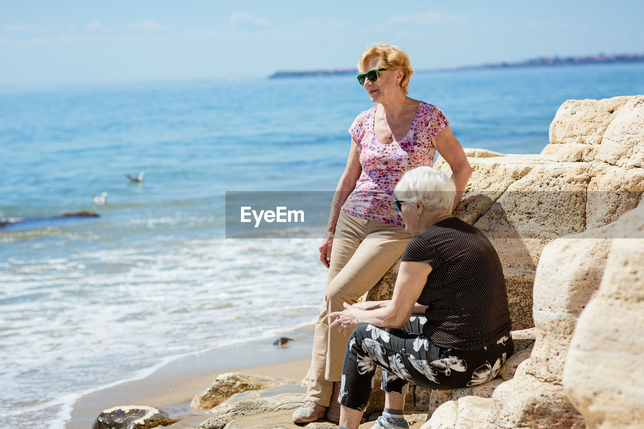 Two elderly women are happy to meet each other, sitting on rock on the seashore