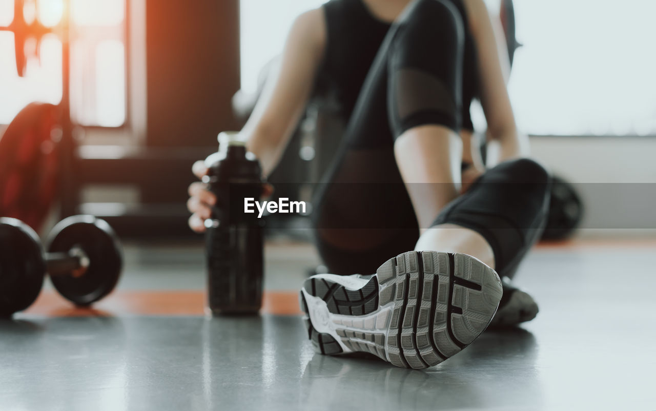 Low section of woman holding water bottle while sitting on floor in gym