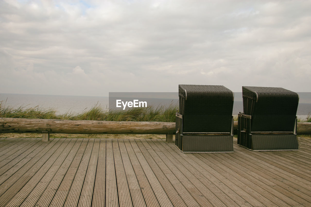 Hooded beach chairs on wooden boardwalk by sea against cloudy sky