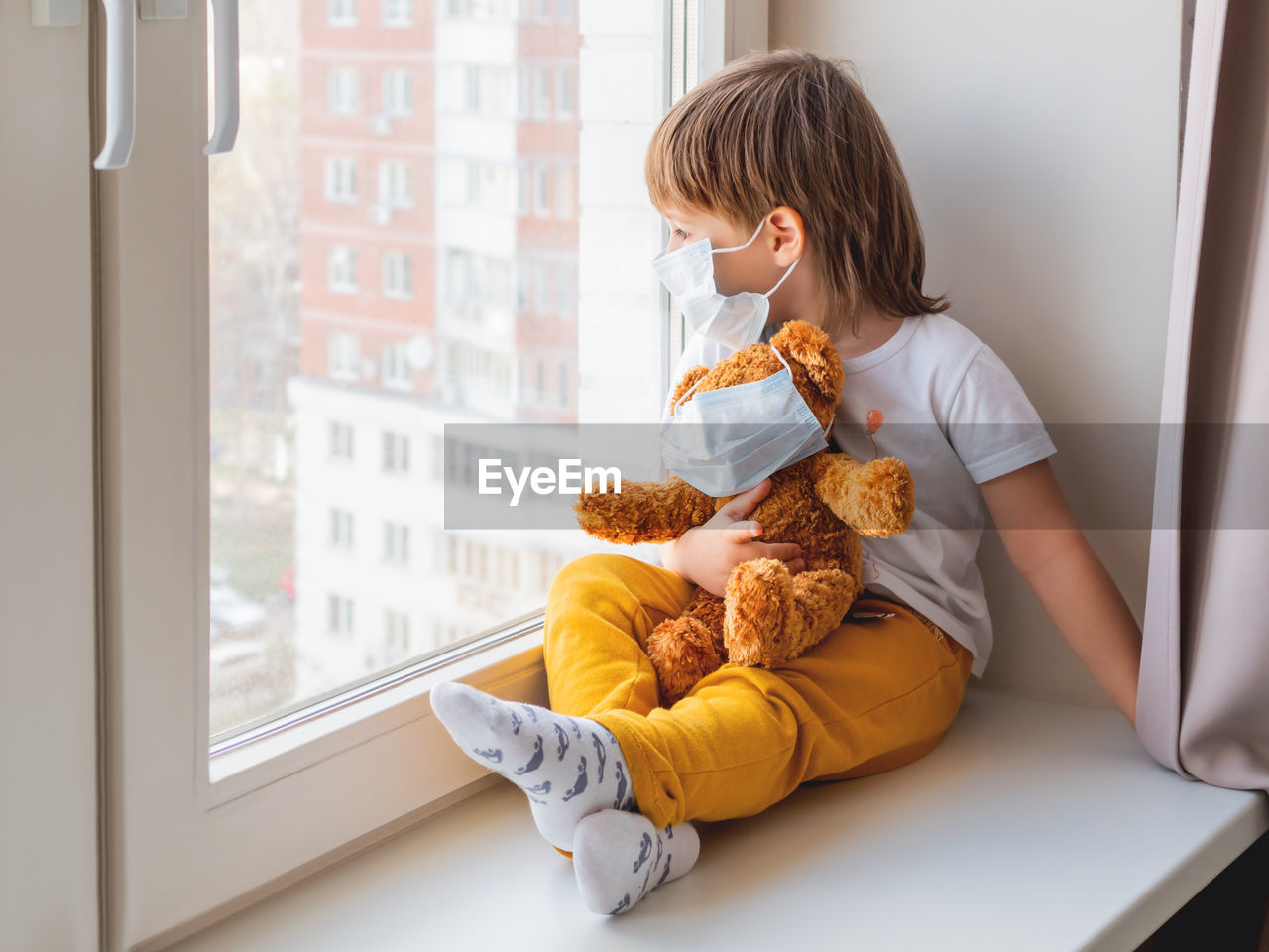 Toddler boy sits with teddy bear in medical masks. kid with plush toy look through window outside. 