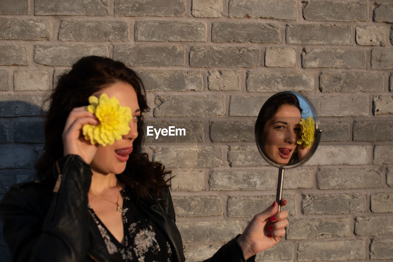 Woman holding yellow flower and mirror with reflection against brick wall