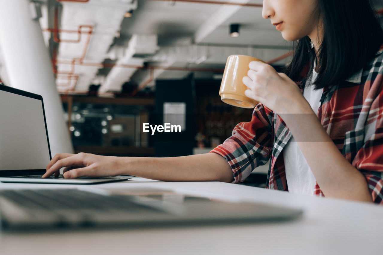 Midsection of woman using laptop while drinking coffee on table