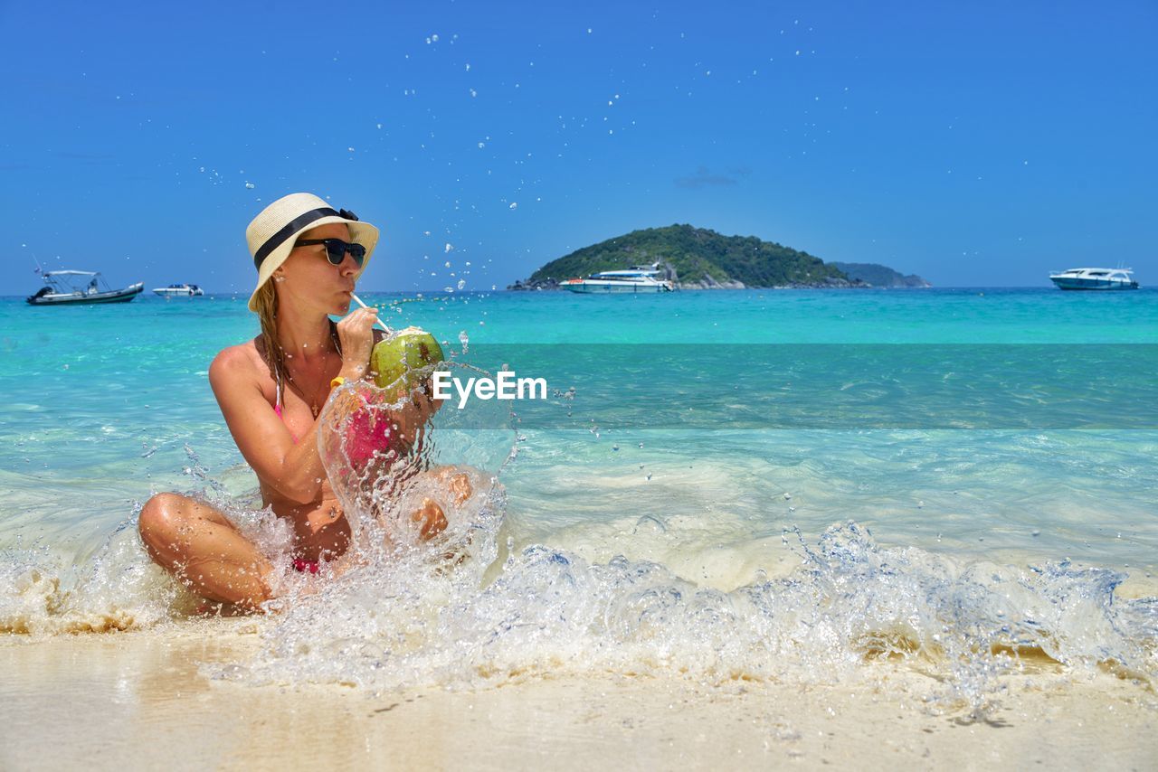 Woman drinking coconut water while sitting at beach against blue sky