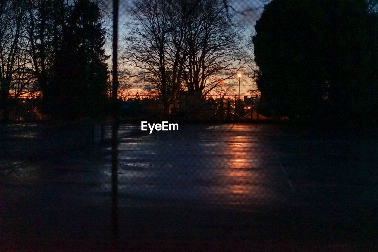 Silhouette trees on field during dusk seen through chainlink fence