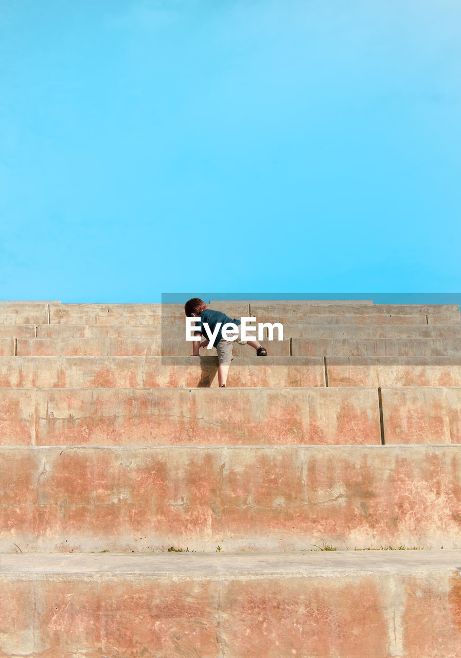 Full length rear view of boy climbing steps against clear blue sky