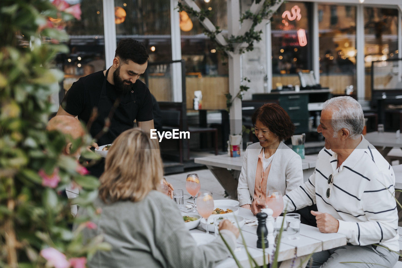 Happy male and female senior friends looking at waiter serving food at restaurant