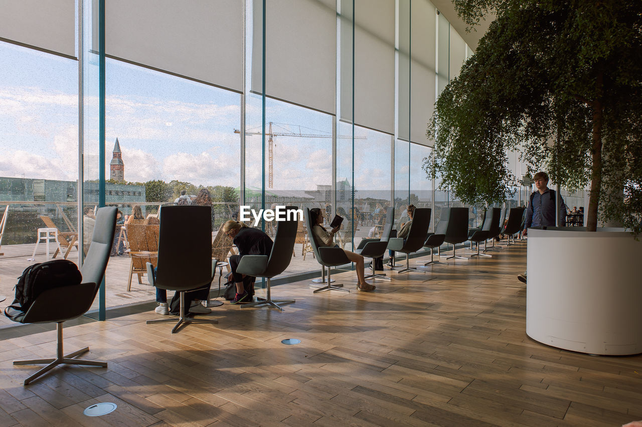 window, chair, business, seat, office, indoors, interior design, glass, table, architecture, adult, day, building, sitting, flooring, nature, furniture, meeting, built structure, business finance and industry, group of people, city, corporate business, men, floor, reflection, sunlight